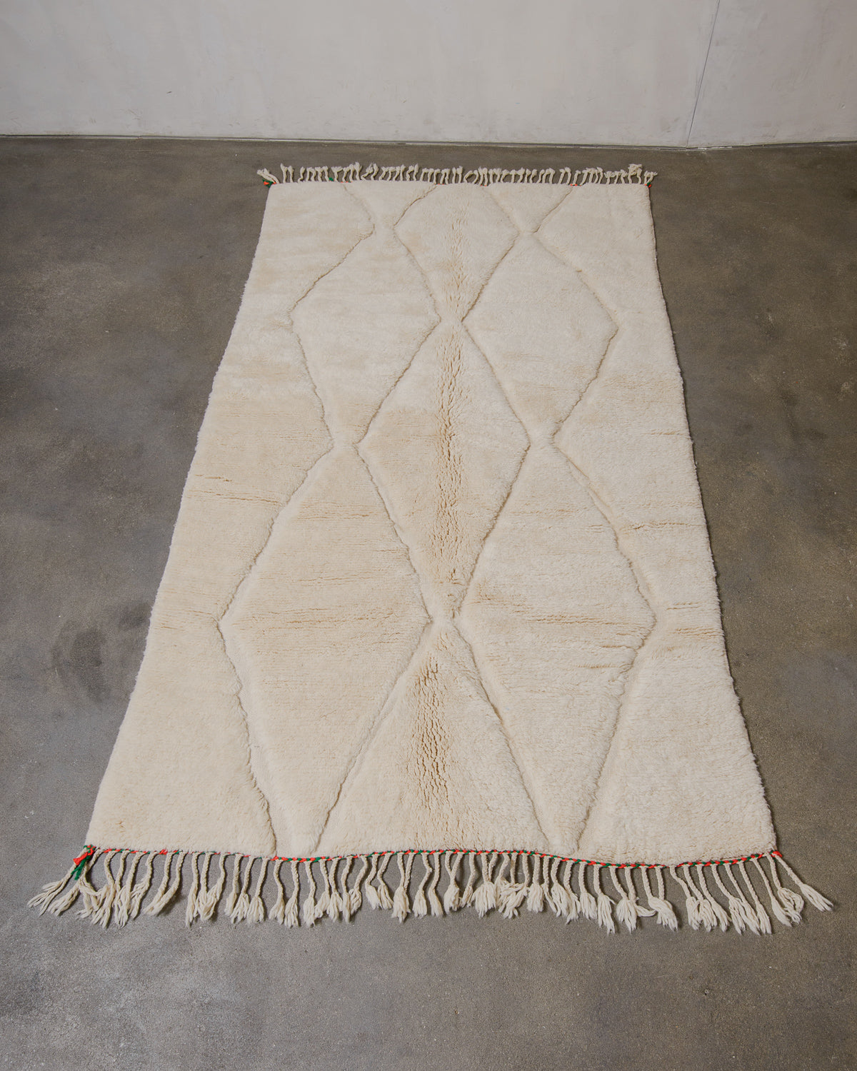 Cream wool rug with geometric pattern and fringes laying on a plain grey floor.
